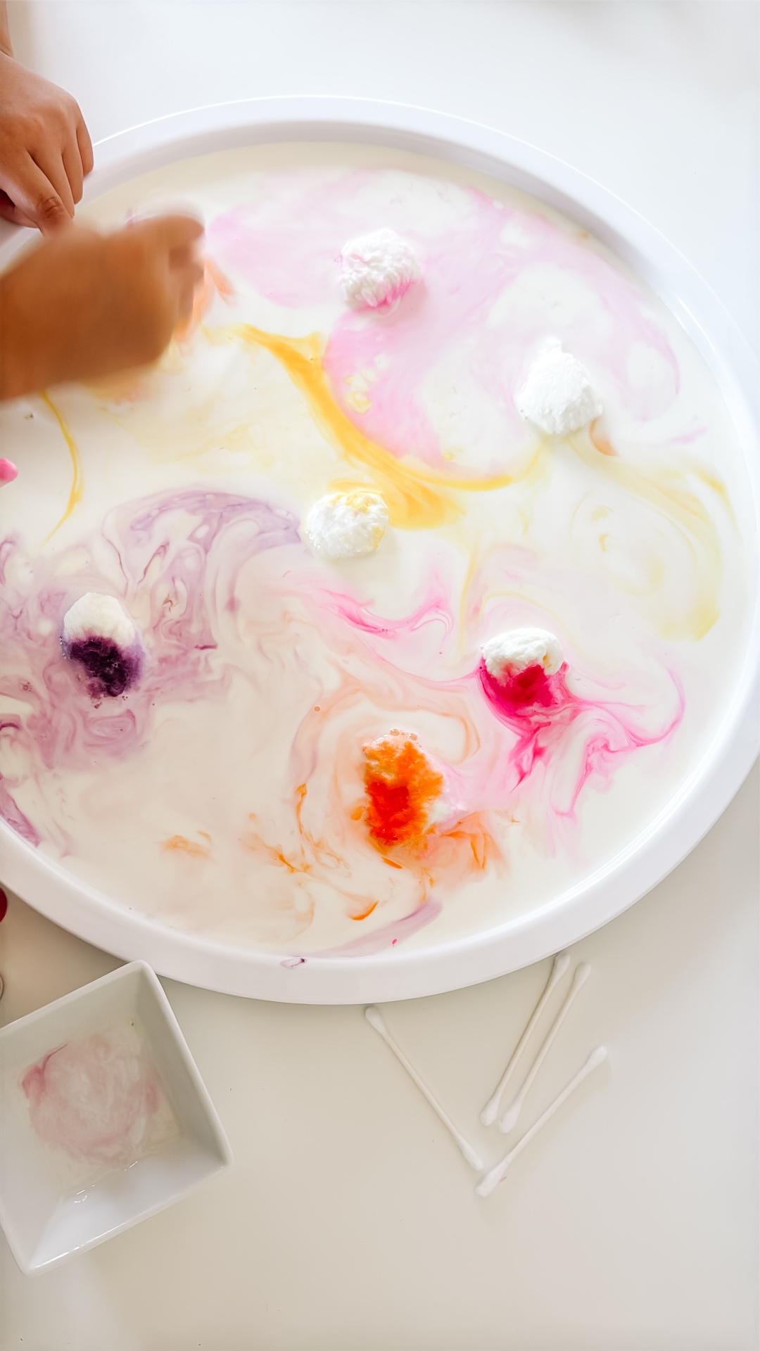 A child using a cotton swab to swirl warm pink, orange, and yellow colors across a tray of milk