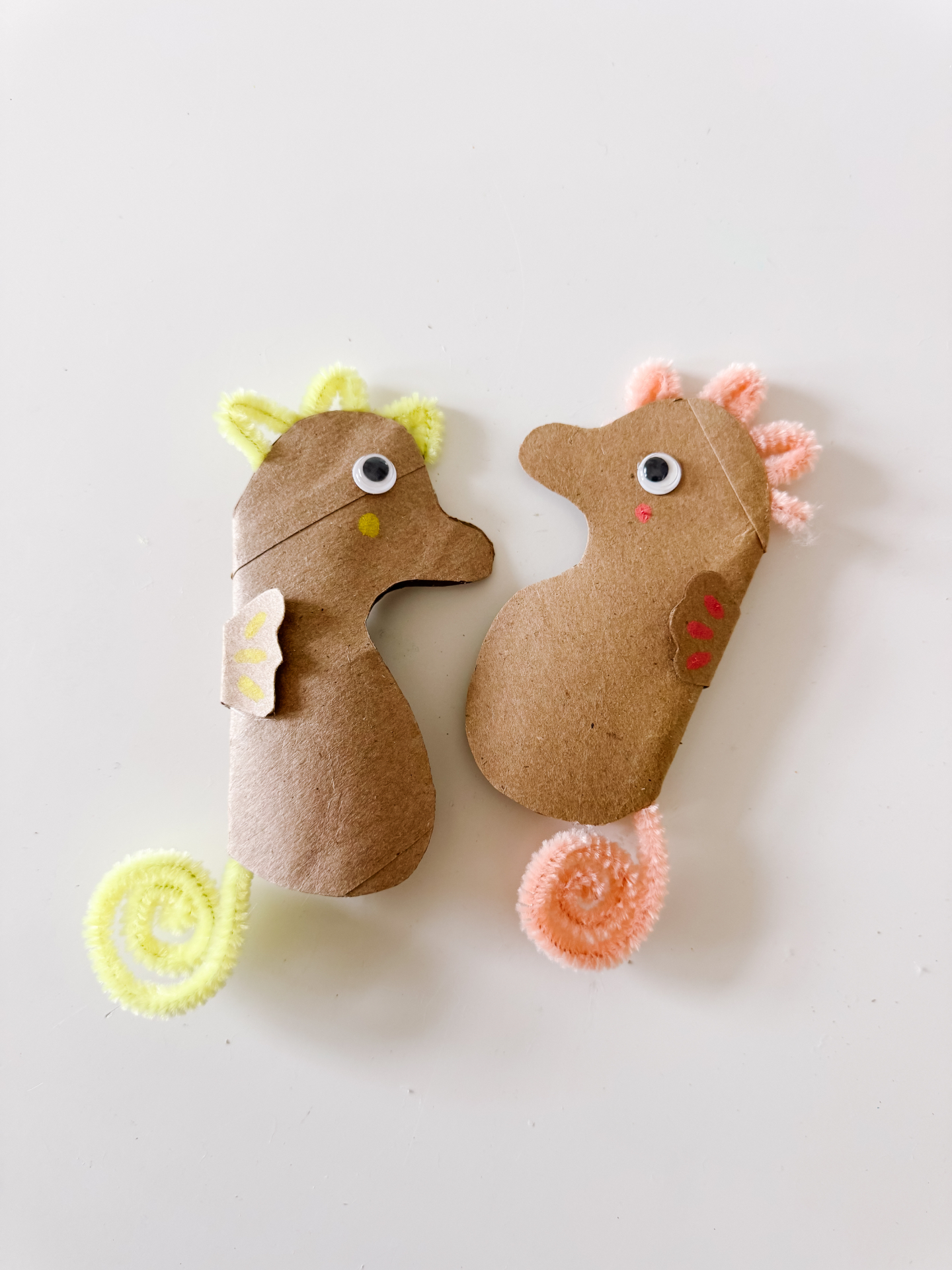 Two flat cardboard seahorses with yellow and pink pipe cleaner tails and fins